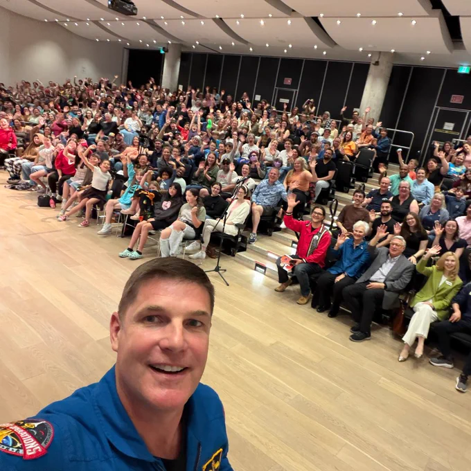 Un astronaute canadien prenant un égoportrait avec un large public dans un auditorium, tout le monde faisant signe à la caméra.