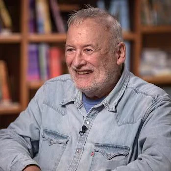 Robert Munsch sits smiling in a light denim button-up shirt, with a small microphone clipped near his chest. He is seated in front of softly blurred bookshelves filled with colourful books, giving the photo a warm library or interview setting.
