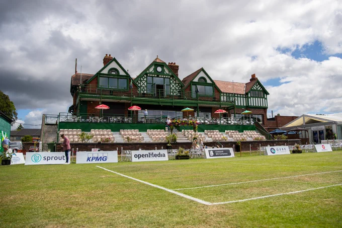 A club house at a cricket ground which has a tennis court set up in front of it. 