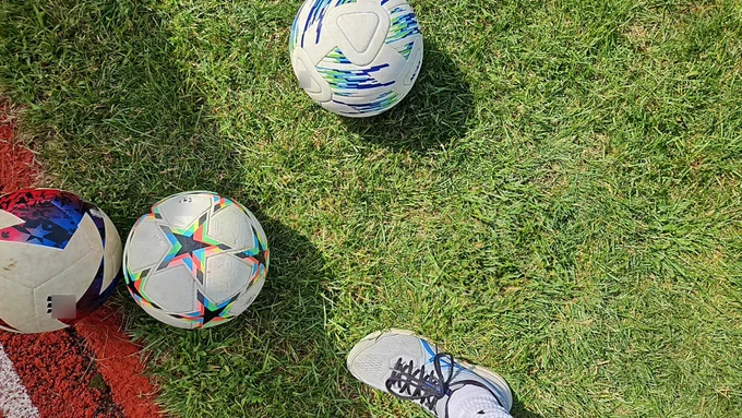 Photograph of three soccer balls on green grass. There is red and white painted concrete visible in the bottom left corner of the image. A person’s foot, clad in a tennis shoe, is visible near the bottom of the photo, indicating that the person is taking a photograph straight down at the ground below their feet. Credit: NASA GLOBE