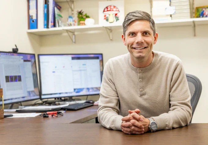 University of South Carolina Floyd School of Medicine researcher Colin Evans poses at his office desk. In the background are two computer monitors with work pulled up on them, and a bookshelf filled with literature and decor.