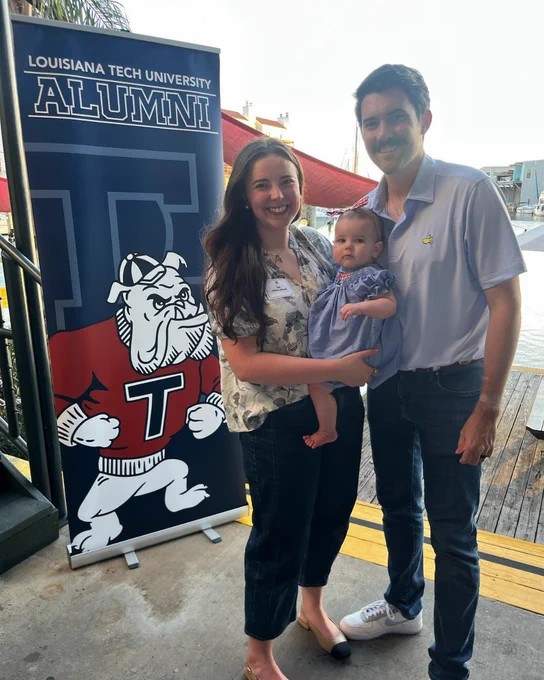 A couple holds their baby while standing in front of a vertical Louisiana Tech alumni banner