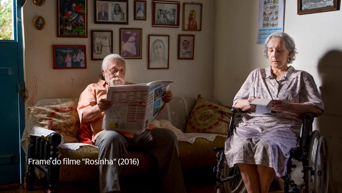 Frame do filme “Rosinha” que destaca um homem idoso sentado em um sofá, segurando um jornal, e uma mulher idosa sentada em uma cadeira de rodas, segurando um papel.