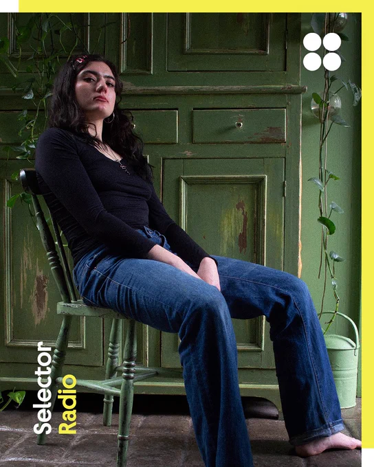 A young dark haired woman, Eaves Wilder, sits casually on a painted wooden chair in front of a weathered green cabinet, surrounded by trailing vines. Their relaxed pose and direct gaze create a calm, confident portrait in a rustic indoor setting.