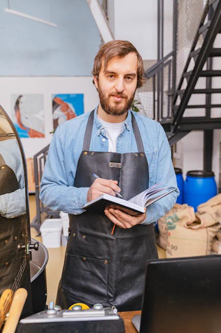 Bearded man in apron holding a notebook and pen inside a coffee shop.