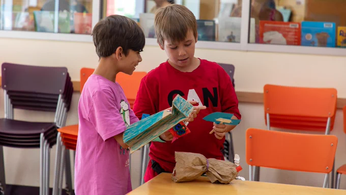 Two kids stand together by a table in the library, curiously examining the paper and stick puppets they have made. The child on the left wears a paper bag puppet coloured blue on their right arm. There are black and orange chairs stacked in the background against the wall.