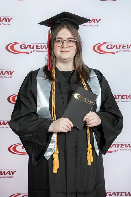 Graduate in black cap and gown holding a Gateway Technical College diploma cover against a branded backdrop.