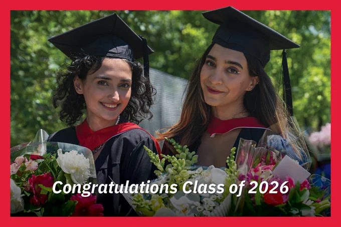 Two graduates in academic regalia smile while holding bouquets. The text reads, "Congratulations Class of 2026." They are outdoors with greenery in the background.