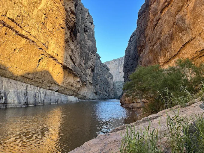 Sunlit canyon walls rise steeply above a calm river in Big Bend. Golden morning light illuminates the rock face on the left while the river winds through the narrow canyon beneath a clear blue sky.