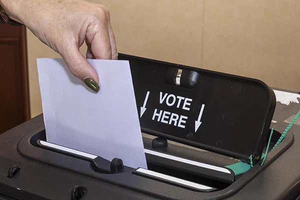 A hand inserts a ballot paper into a ballot box labelled "VOTE HERE".