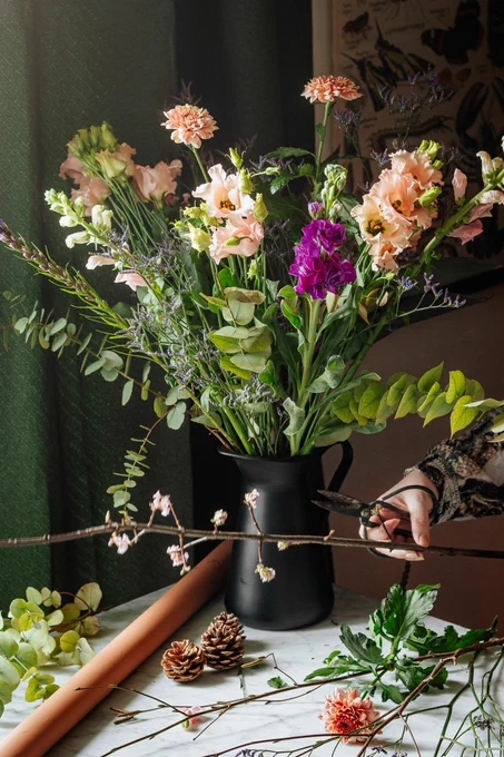 A bouquet of flowers in a black jug with an off camera person holding scissors and flowers in hand. On the table is a roll of paper, pinecones and various foliage. 