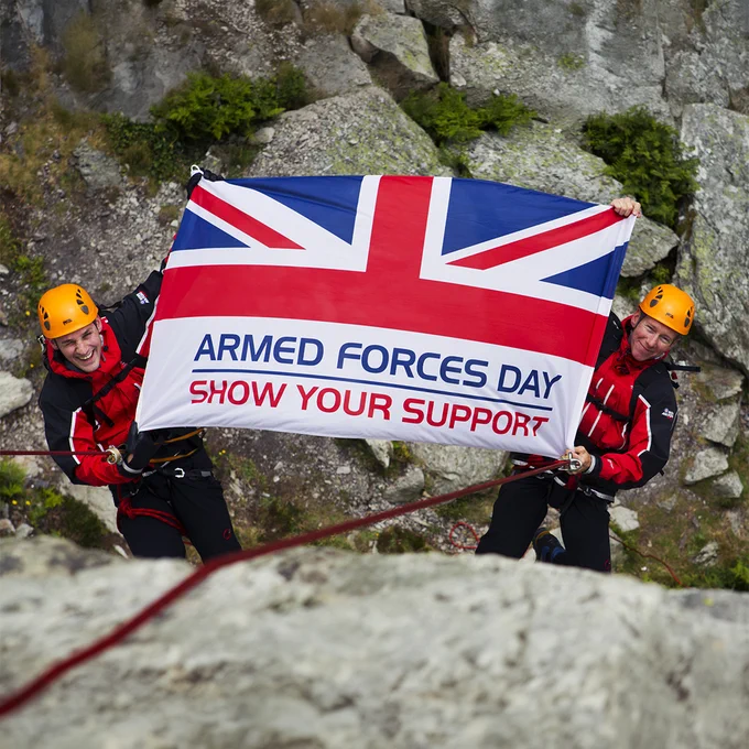 Two climbers in red and black gear and orange helmets hang on ropes against a rocky cliff, holding a large Union Jack banner that reads, “Armed Forces Day – Show Your Support.”