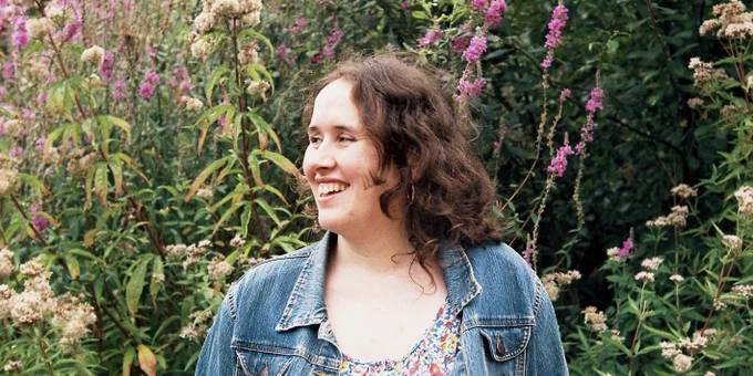 RPS Composer Emily Hazrati looking to the left, wearing a denim jacket with flowers and foliage behind her.