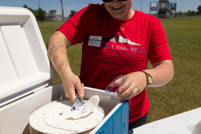A person wearing a red "The Wall That Heals" T-shirt is scooping ice cream from a large container at an outdoor event. The shirt features sponsors like the Vietnam Veterans Memorial Fund. The person has a tattoo on their arm and sunglasses on.