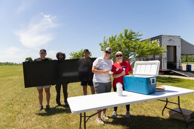 A group of people stand outdoors on a sunny day, near a white table with a blue cooler. One person holds a large black panel, partially obscuring the group. A few trees and a truck are visible in the background.