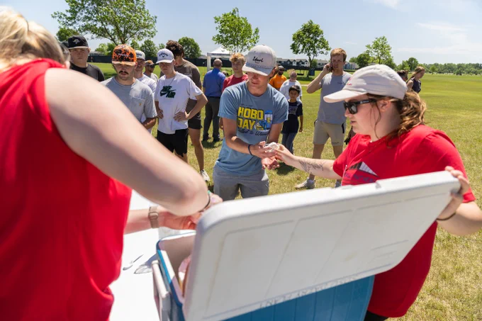 A group of people outdoors on a grassy field, gathered around a cooler. Some individuals are distributing ice cream. One person is wearing a shirt with the words "Hobo Day." Trees and a clear blue sky are in the background.