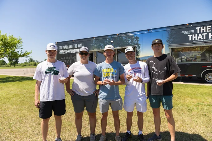 A group of five people standing on grass, holding dishes of ice cream and smiling. Behind them is a trailer with "The War and the Wall" written on it, under a clear blue sky.