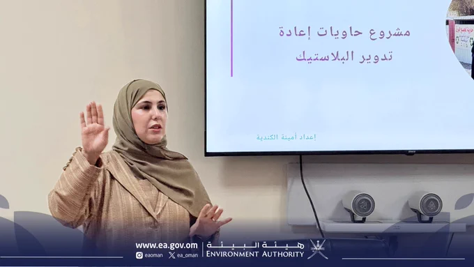 صورة فتاة تقدم عرضاً مرئياً وخلفها شاشة تظهر نص ( مشروع حاويات إعادة تدوير البلاستيك)