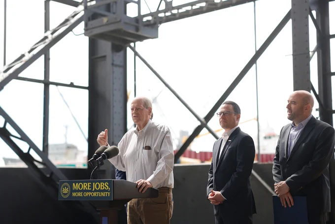 Dick Hayne, CEO, Urban Outfitters speaking from a podium, addressing an audience at a podium with a sign reading 'More Jobs, More Opportunity' inside an industrial setting.