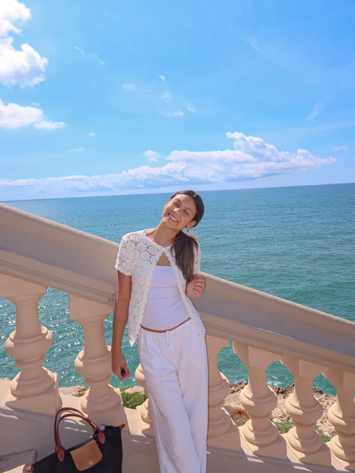 A person is smiling and leaning against a stone balustrade with an ocean view in the background. The sky is bright blue with fluffy clouds. A black and brown bag is placed nearby.