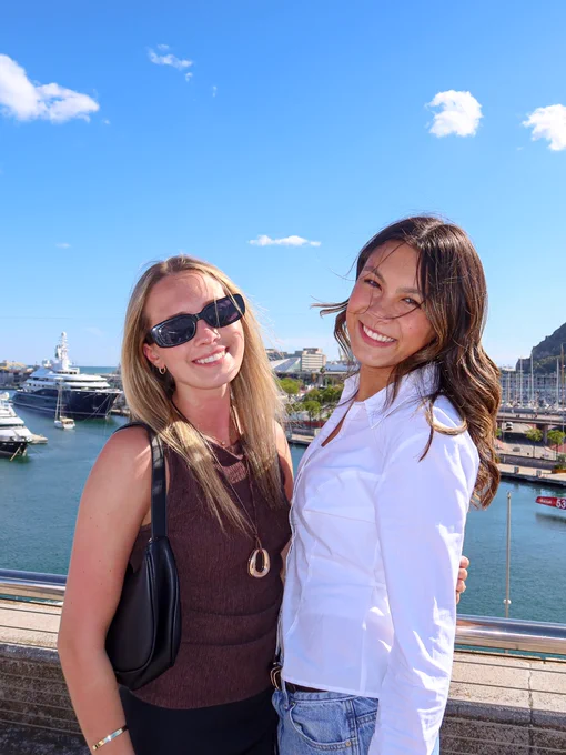Two students posing together with a marina in the background on a sunny day.