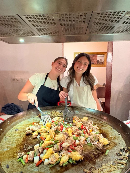 Two people smiling and cooking on a large round griddle filled with colorful ingredients like vegetables and pieces of meat. One person is holding a spatula and is wearing an apron. The setting is a kitchen with cooking appliances above them.