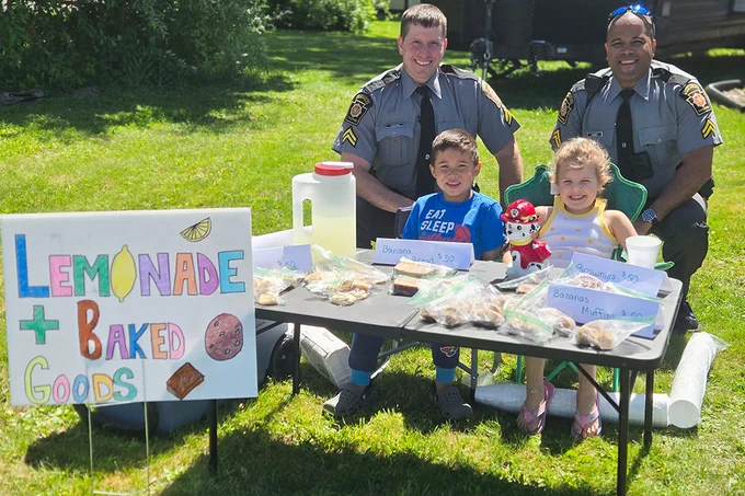 On a sunny day in a grassy yard, two Pennsylvania State Police Corporals kneel behind two children sitting at a table with lemonade and baked goods on display