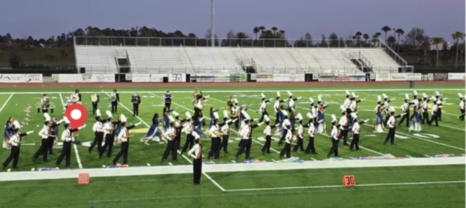 A large marching band performs on a football field during the evening, with musicians and color guard members in uniform. The stands in the background are mostly empty, adding a calm atmosphere to the scene.