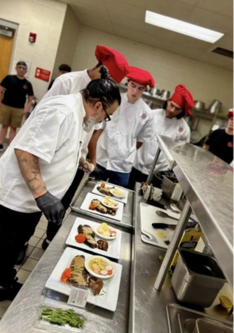 Chefs in white uniforms with red hats examine neatly plated dishes in a professional kitchen, displaying focus and teamwork.