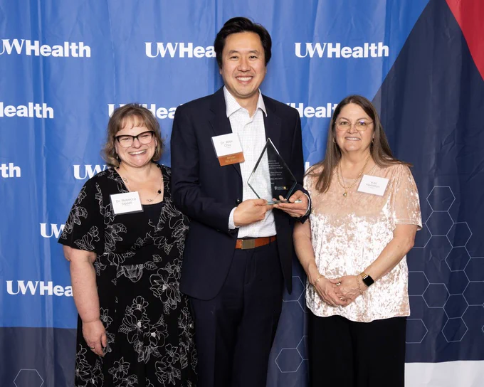 Dr. Rebecca Sippel, Dr. Alex Chiu, and Kar Hamilton stand together in front of a banner that says UW Health. Dr. Chiu is holding an award.