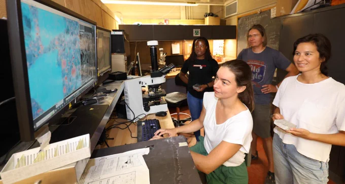 Four researchers in a lab around a microscope and computer station. From left: Wisdom Okoh, Katia Ossetchkina seated at the keyboard, Professor Karl Peterson and Amy Montgomery, who holds a concrete sample. A monitor shows a magnified microscopic image of concrete. 