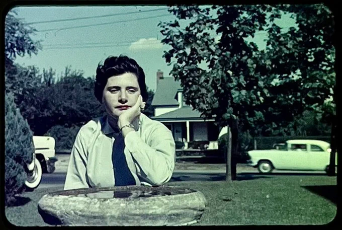 Nancy Walker rests her head on a stone birdbath. Behind her is a house and car. 