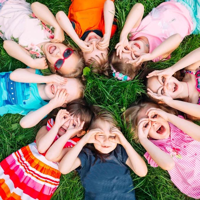 Children lying on green grass in a circle, wearing bright, colourful summer clothes. Each child has their head close together and is holding their hands up to their faces, making playful shapes around their eyes. The scene is outdoors in daylight with vibrant colours and a fun, energetic feel.