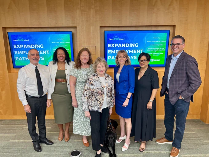 A group photo with 7 adults smiling for the camera in front of screens that show the text: "Expanding Employment Pathways".