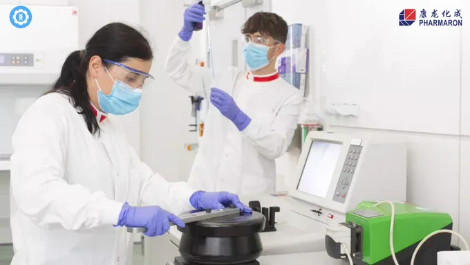 Advancing Gene Therapy Innovation. Image of two scientists in a lab setting, one using a pipette and the other operating a centrifuge. Both are in white lab coats, blue gloves, and face masks, highlighting the professional research environment at Pharmaron