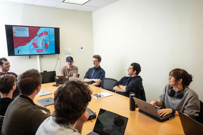 Eight Mason Student Government students sit around a wooden conference table, each with a laptop or notebook open as they discuss a project. On the wall behind them, a large screen displays a colorful map titled “Arctic Mission 2019,” showing red and blue labeled zones across land and ocean. The group looks focused and collaborative, capturing a moment from an SG meeting or planning session.