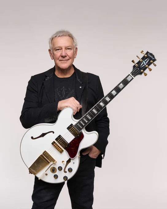 Alex Lifeson from the band Rush stands holding a white Epiphone signature Inspired by Gibson Custom 1976 ES-355 guitar with gold hardware against a plain background. The person is wearing a dark jacket and shirt.