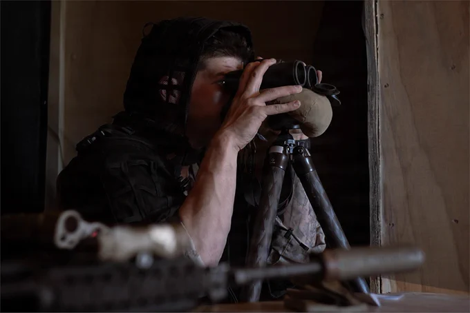 A Marine observes terrain through binoculars from an observation position inside a building.
