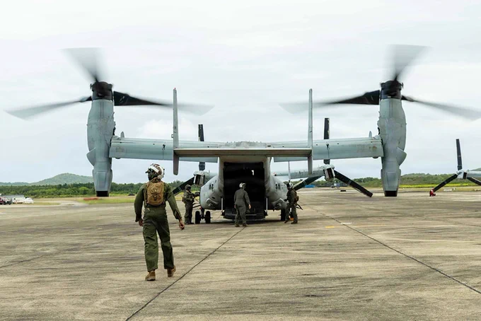 A Marine approaches a MV-22B Osprey with its rear ramp lowered during aviation operations on a tropical airfield.