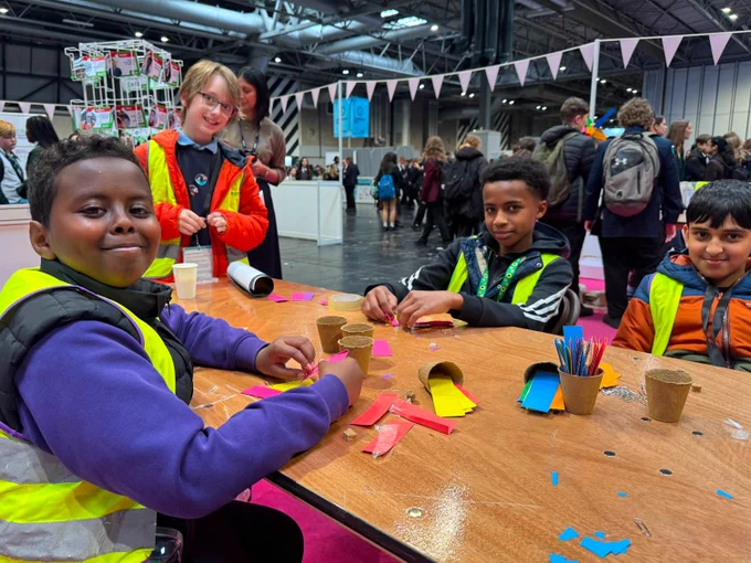 Children sit at a table engaging in a craft activity, using colorful paper and stickers. They are in a large indoor venue filled with other visitors. The atmosphere is lively and creative.