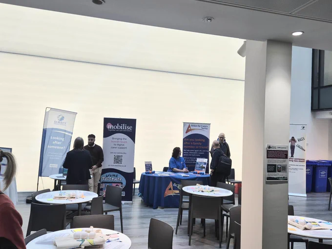 Attendees speaking with organisations at information stands during a carers event at Victoria Gate, Woking, featuring banners from Surrey County Council, Mobilise, and Action for Carers Surrey.
