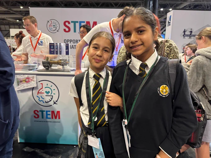 Students in school uniforms stand together at a STEM exhibition booth. The background includes displays related to science, technology, engineering, and mathematics, with visible STEM logos and informational materials. Other attendees are in the background engaging with the exhibit.