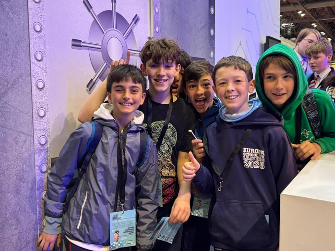 A group of young people smiling and posing in front of a large, metallic vault door at an indoor event. Some of the individuals are wearing lanyards. The background has industrial design elements and there are other people visible in the distance.
