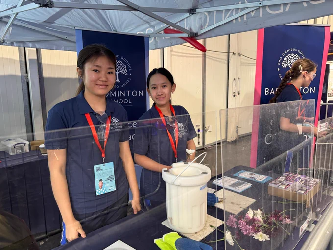 Two people standing behind a table in a Badminton School stand. They are wearing blue shirts with name tags and are under a canopy with the School logo. The table has various items including a bucket, flowers, and pamphlets.