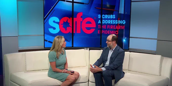 A screengrab of Dr. James Bigham on WMTV 15 news, sitting on a couch with a reporter in front of a screen that reads "Safe: scrubs addressing the firearm epidemic."