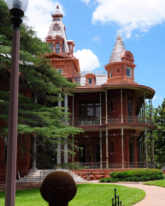 Ornate red brick mansion under a blue sky, featuring multiple towers, one conical and one spire-like, with intricate iron balconies. A white staircase leads to the entrance, flanked by green trees and a lamppost on the left. Green lawn with a paved path in the foreground.