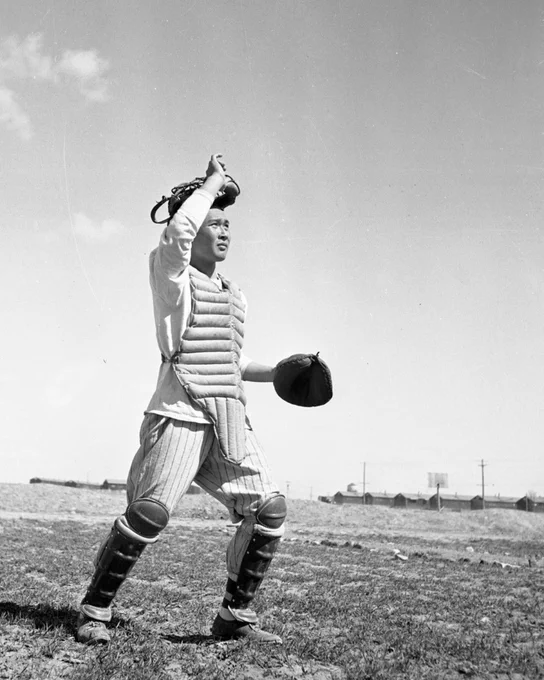 Black-and-white photo of a young Japanese American man in full baseball catcher's gear — chest protector, shin guards, and mitt — looking into the distance. He stands on an open dirt field with barracks visible in the distant background.