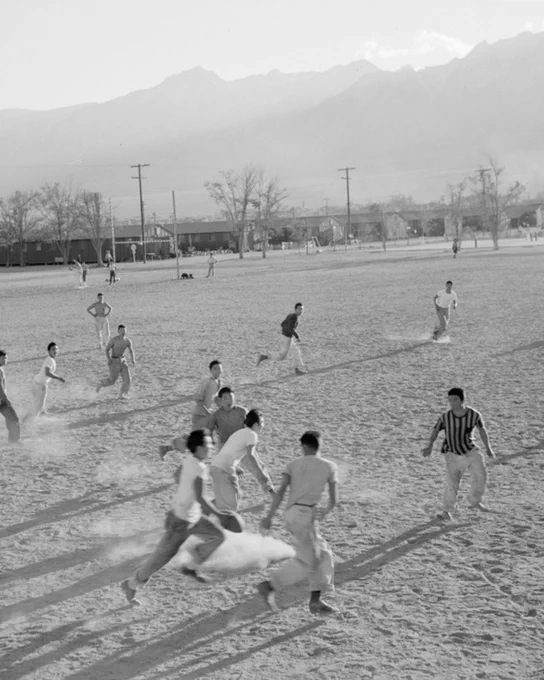 Black-and-white photo of a group of Japanese American men playing football on a large dirt field. Mountains are visible in the background, along with barracks. Players are mid-action, running and tackling.