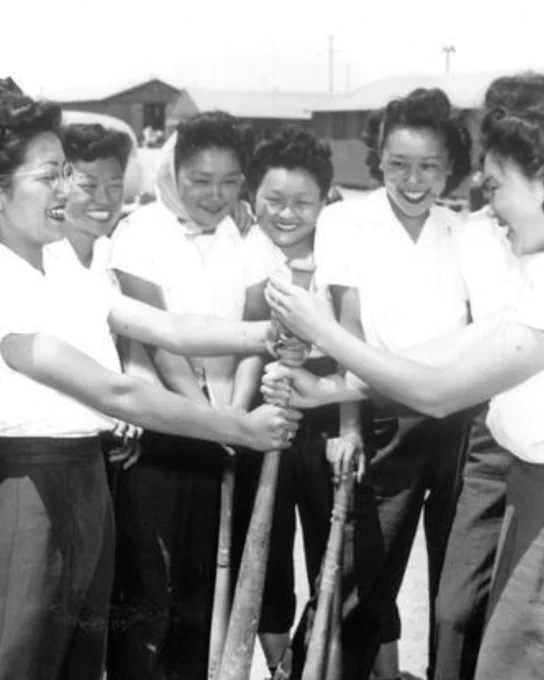 Black-and-white photo of six smiling Japanese American women in white shirts, gathered together and gripping a baseball bat in a classic "pick teams" hand-over-hand stacking pose. Barracks are faintly visible in the background.