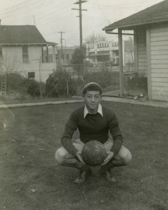 Black-and-white photo of a young Nisei boy crouching in a backyard, holding a round ball with both hands. He wears a dark long-sleeve sweater over a collared shirt and shorts. Residential houses and utility poles are visible in the background.